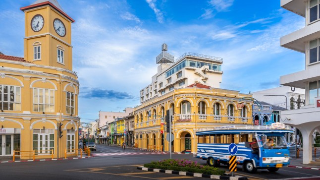 Phuket, Thailand - 11 November 2024: Menara Jam Kuning yang ikonik dan gedung Chartered Bank yang bersejarah di Kota Tua Phuket, menampilkan arsitektur Tiongkok-Portugis, membingkai persimpangan yang sibuk dengan bus lokal berwarna biru yang lewat di sana
