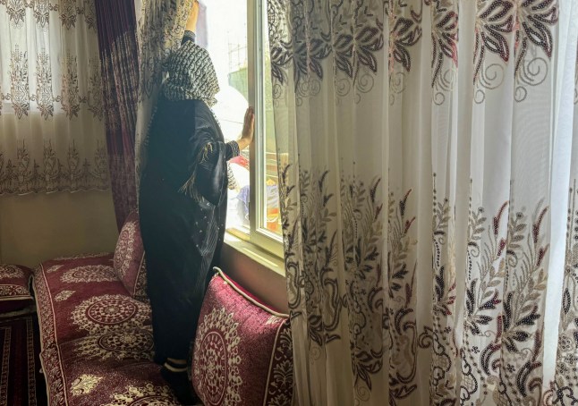 Dalam foto yang diambil pada 3 April 2024 ini, Shirin, yang sedang mengikuti program master online, menarik tirai jendela saat wawancara dengan AFP di Kabul. Pada bulan Desember 2021, pemerintah Taliban mewajibkan setiap wanita yang melakukan perjalanan lebih dari 72 kilometer (45 mil) untuk didampingi oleh mahram. "Banyak wanita tidak memiliki pria di rumah," kata Shirin, 25, yang sedang belajar untuk mendapatkan gelar master secara online setelah dilarang masuk universitas oleh pihak berwenang. Tahun lalu, Shirin mengatakan pihak berwenang Taliban mengganggu acara piknik karena dia dan anggota keluarga perempuan lainnya berada di dalam minibus yang dikemudikan oleh sepupu mereka – satu-satunya laki-laki, bukan mahram. (Foto oleh AFP) (Foto oleh -/AFP via Getty Images)